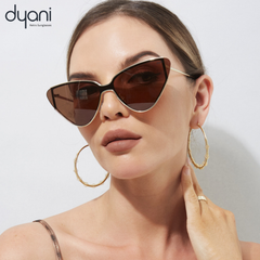 Gold-framed cat-eye sunglasses with dark lenses by Dyani on a white background, worn by a model with hoop earrings.