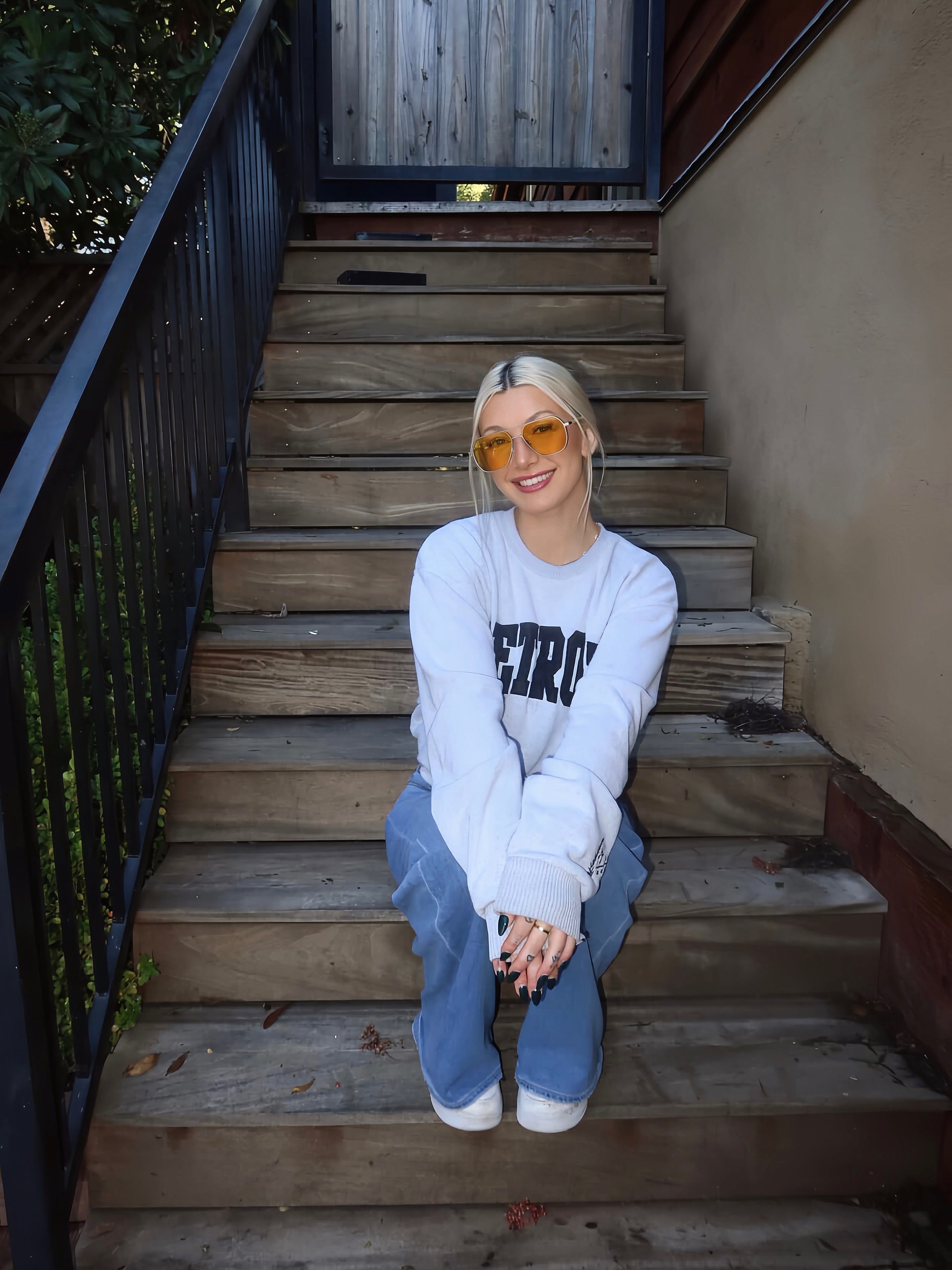 Person sitting on wooden steps wearing a white sweatshirt with text and blue jeans.